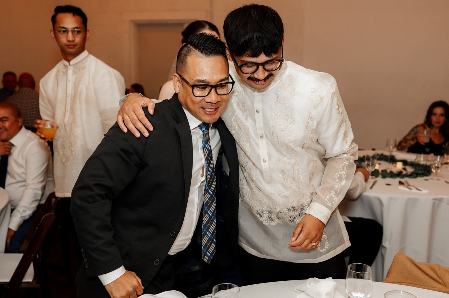 Two men sharing a warm embrace, one wearing a suit and the other in a Barong Tagalog, surrounded by the sophisticated setting of a wedding at Union/Pine.