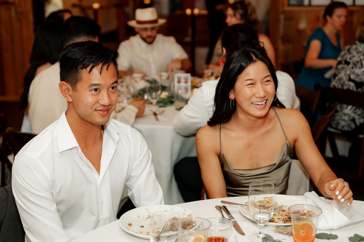 Two people smiling at a dining table with plates and drinks, surrounded by others in a warmly lit room, capturing the joy of a wedding at Union/Pine.