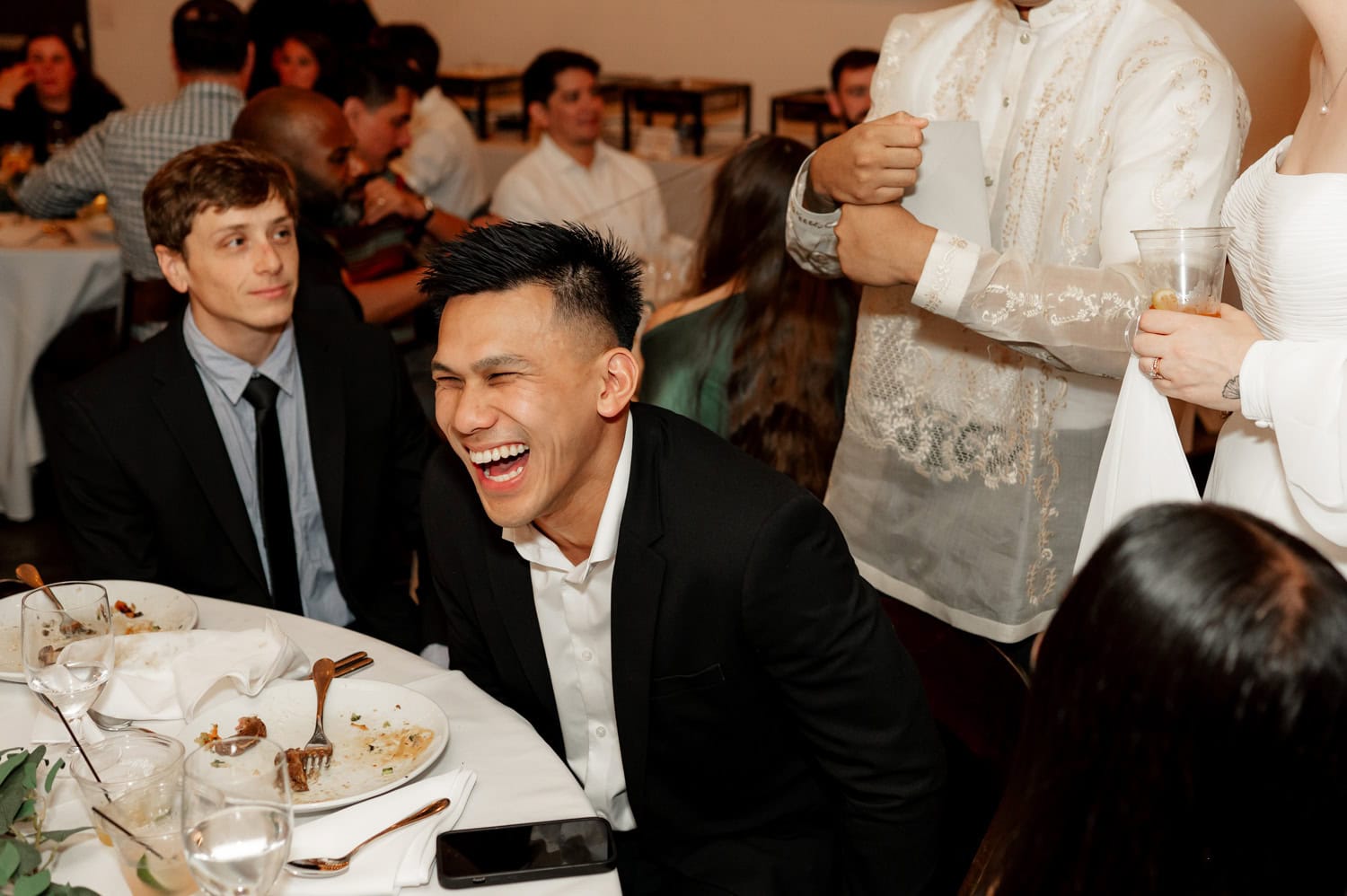 A man in a suit laughs while sitting at a table with others during a wedding at Union/Pine, surrounded by the sounds of clinking glasses and plates of elegantly arranged food.