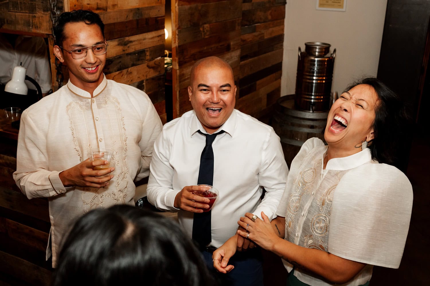 Three people laughing and holding drinks at a social gathering, dressed in semi-formal attire, celebrating joyfully at a wedding at Union/Pine.