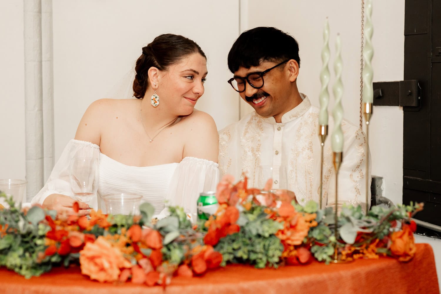 A couple sits at a decorated table, smiling fondly at each other with orange flowers and green leaves in the foreground during their wedding at Union/Pine.