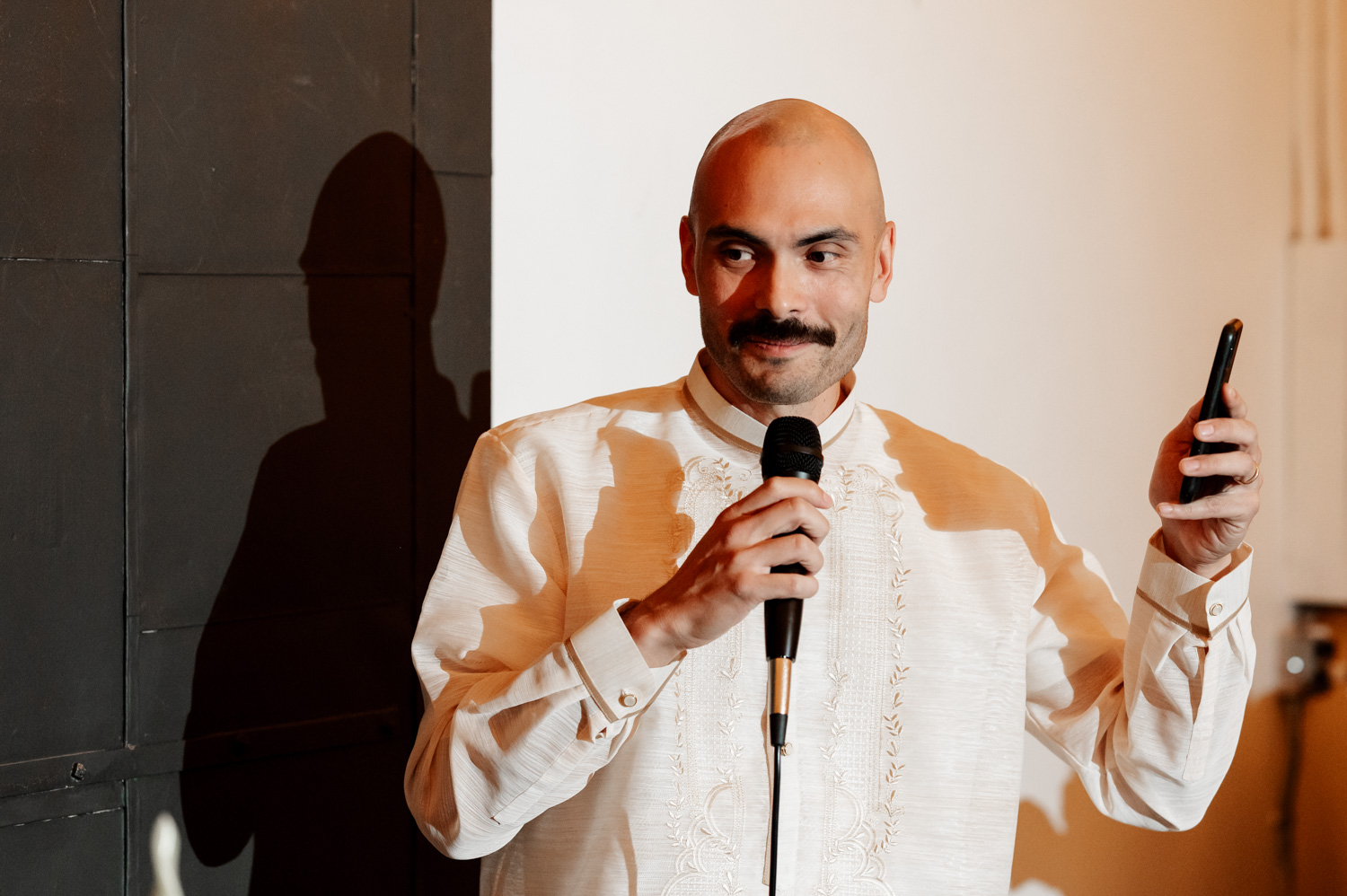 A man in a white embroidered shirt speaks into a microphone at a wedding, holding his phone gracefully. The setting is Union/Pine, and the atmosphere is lively and celebratory.