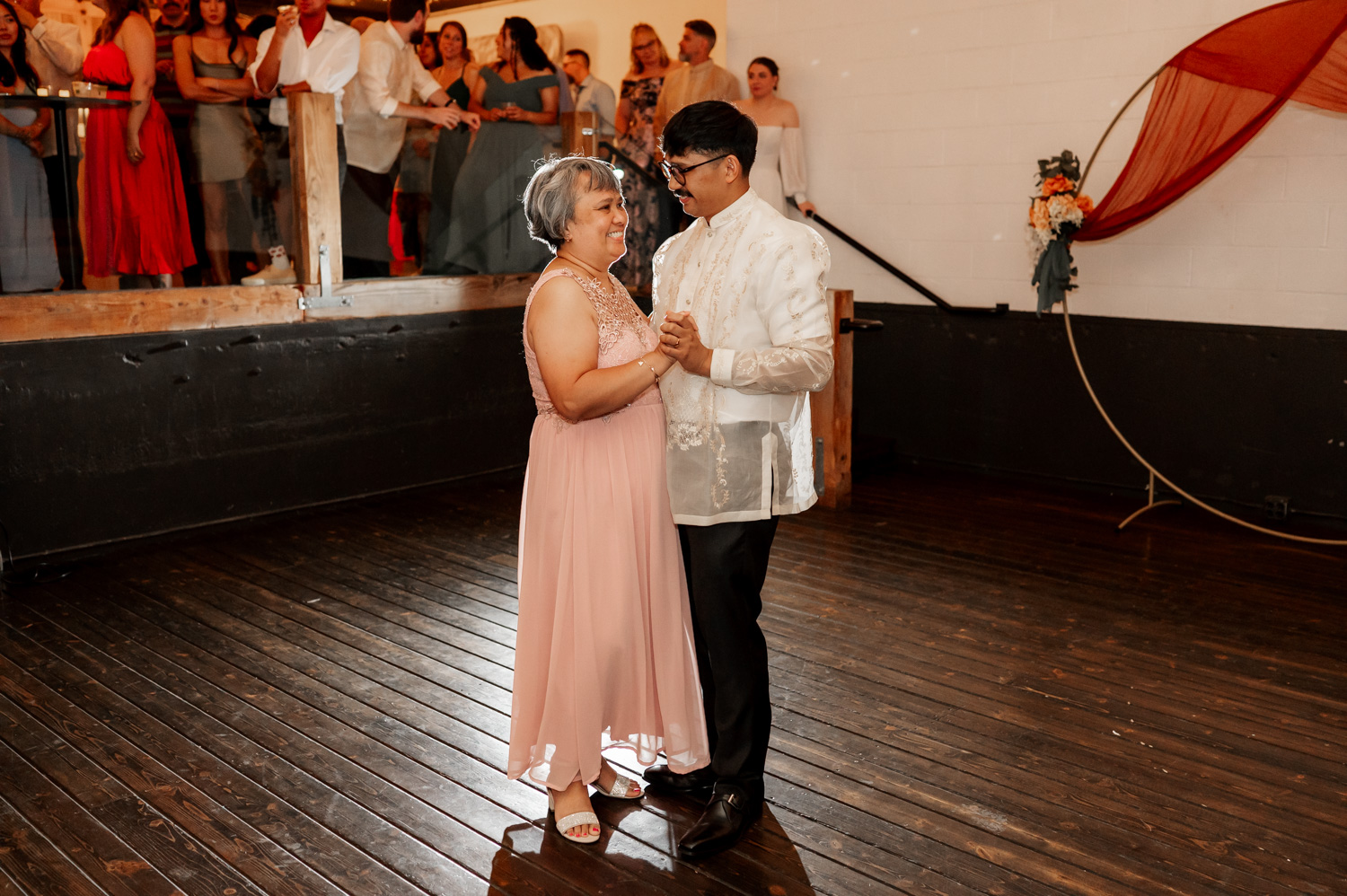 During a wedding celebration at Union/Pine, two people dance joyfully on a wooden floor while an audience watches with admiration.