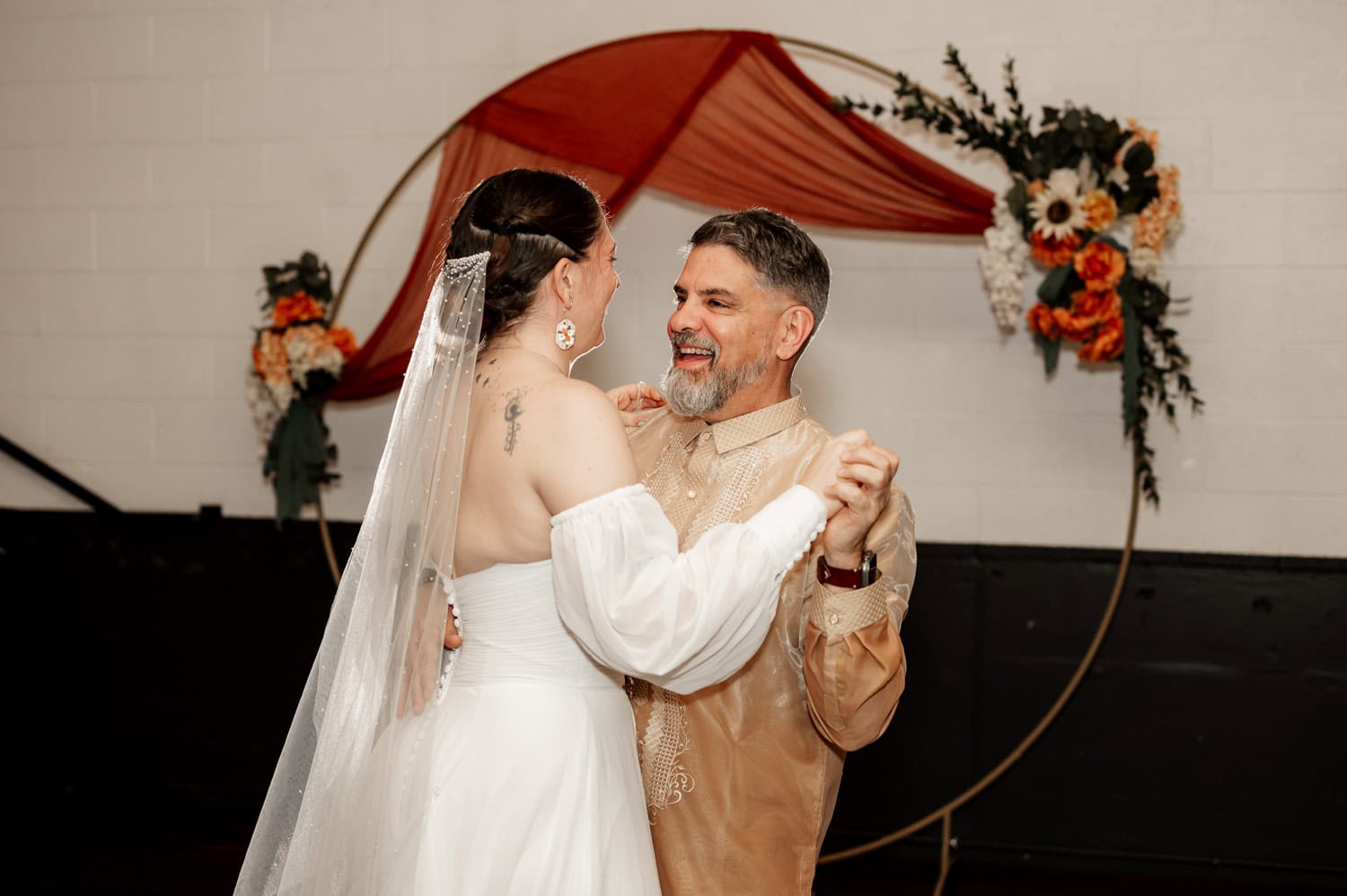 A bride and an older man smile as they dance together in front of a floral hoop backdrop during a wedding celebration at Union/Pine.