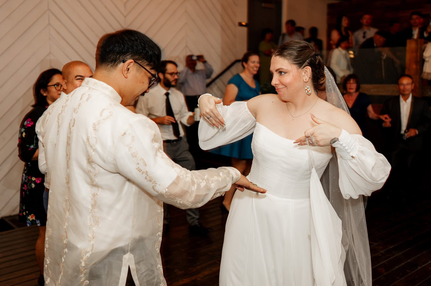 The bride and groom dance joyfully on the wooden floor at Union/Pine, surrounded by smiling wedding guests.