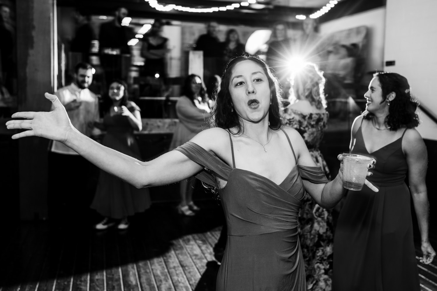 A woman in a dress dances energetically at a lively wedding at Union/Pine, holding her drink tightly. Guests surrounding her are joyfully mingling, contributing to the vibrant atmosphere of the celebration.