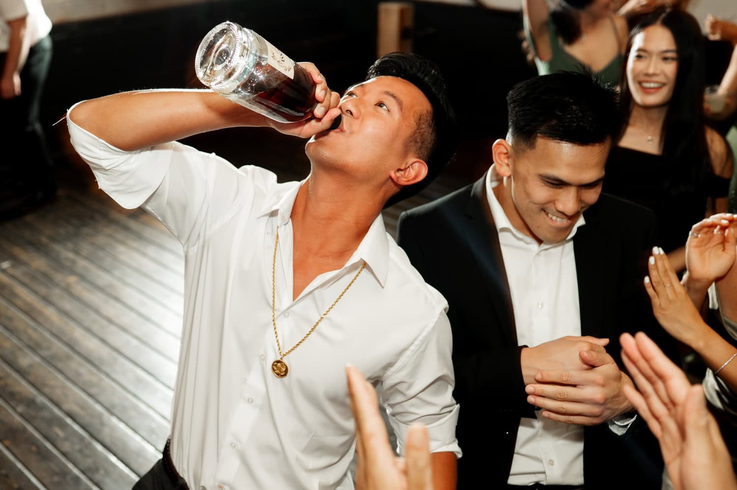 A man in a white shirt drinks from a bottle as people around him cheer and clap at a wedding in Union/Pine, creating an energetic scene.