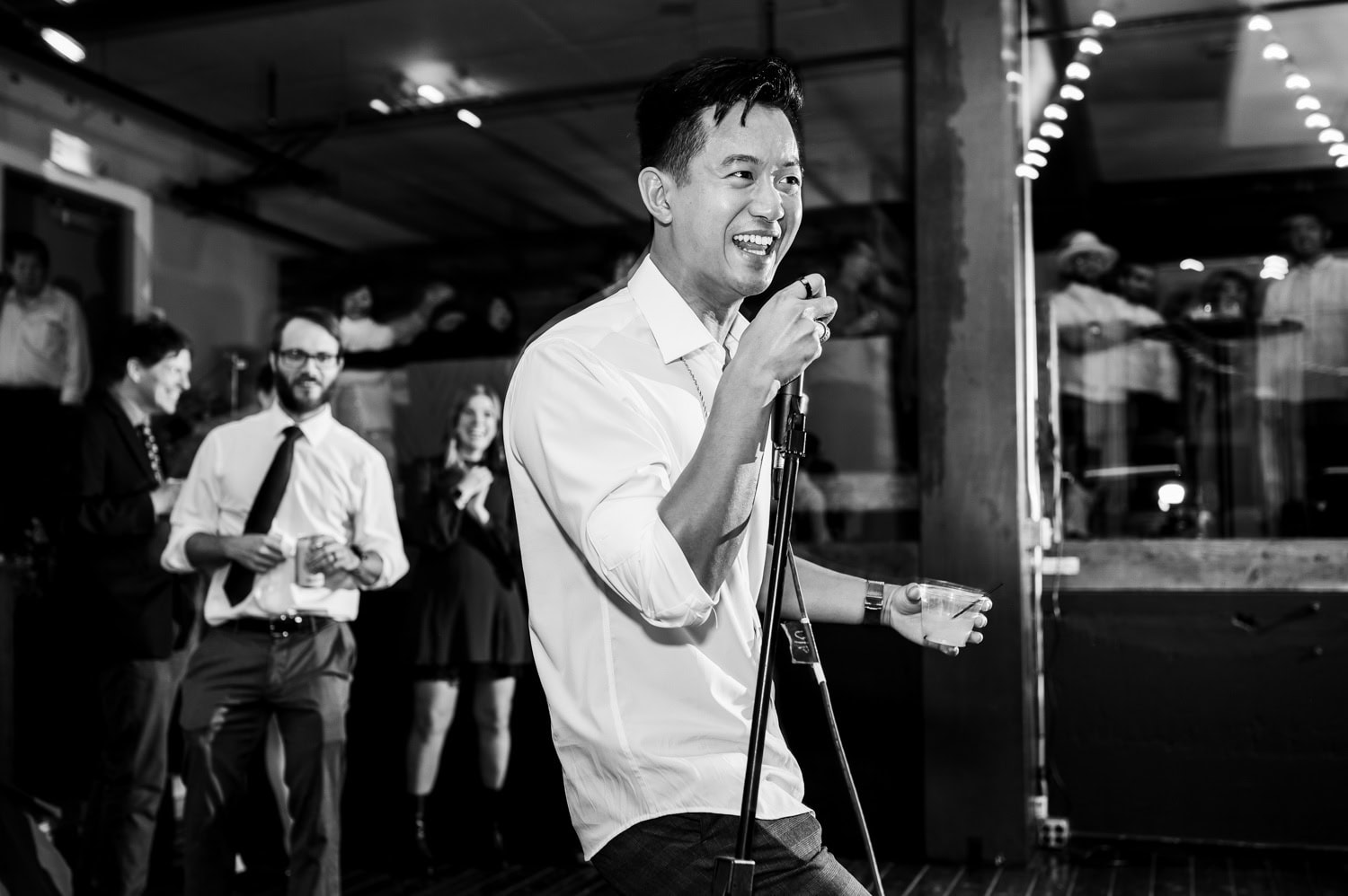 A man sings passionately into a microphone at a wedding in Union/Pine, with guests clapping and smiling in the background.