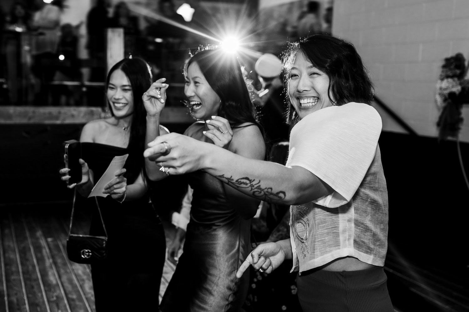 Three women laughing and pointing at an event, one holding a camera. Black and white photo capturing a lively wedding atmosphere at Union/Pine.