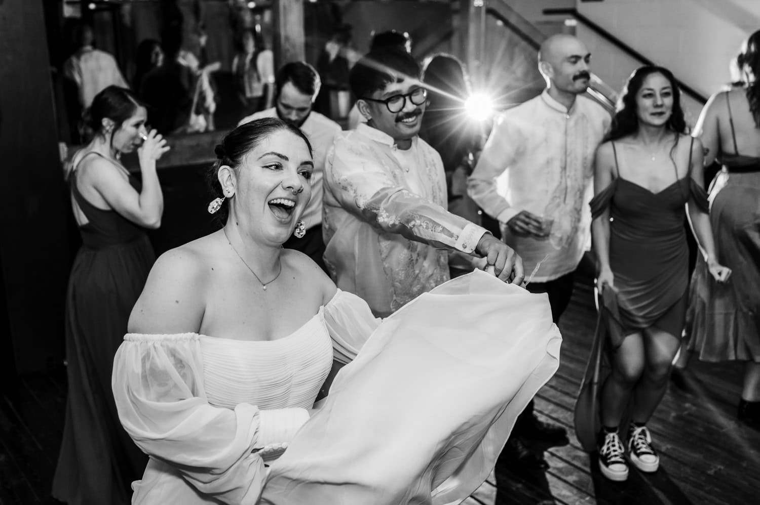 A woman in a white dress dances joyfully with others at a wedding held indoors at Union/Pine. A bright flash highlights the spirited celebration and lively atmosphere, capturing the moment.