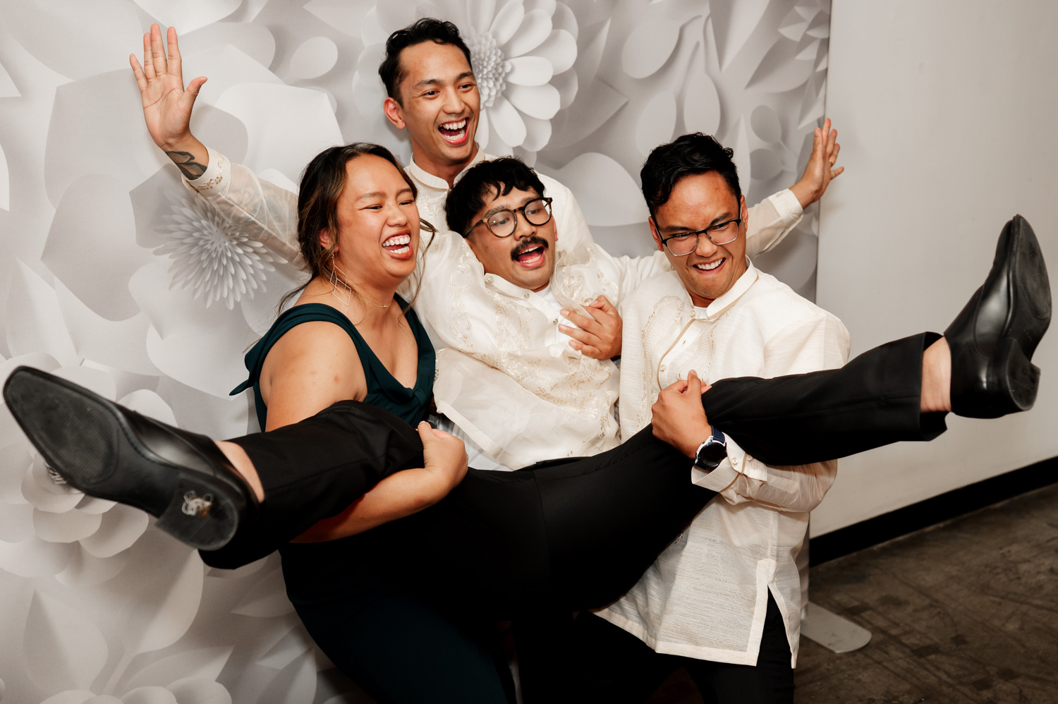 Five people celebrate joyfully in front of a floral-patterned wall. Four individuals hold up another person, all sharing laughs and smiles at Union/Pine.