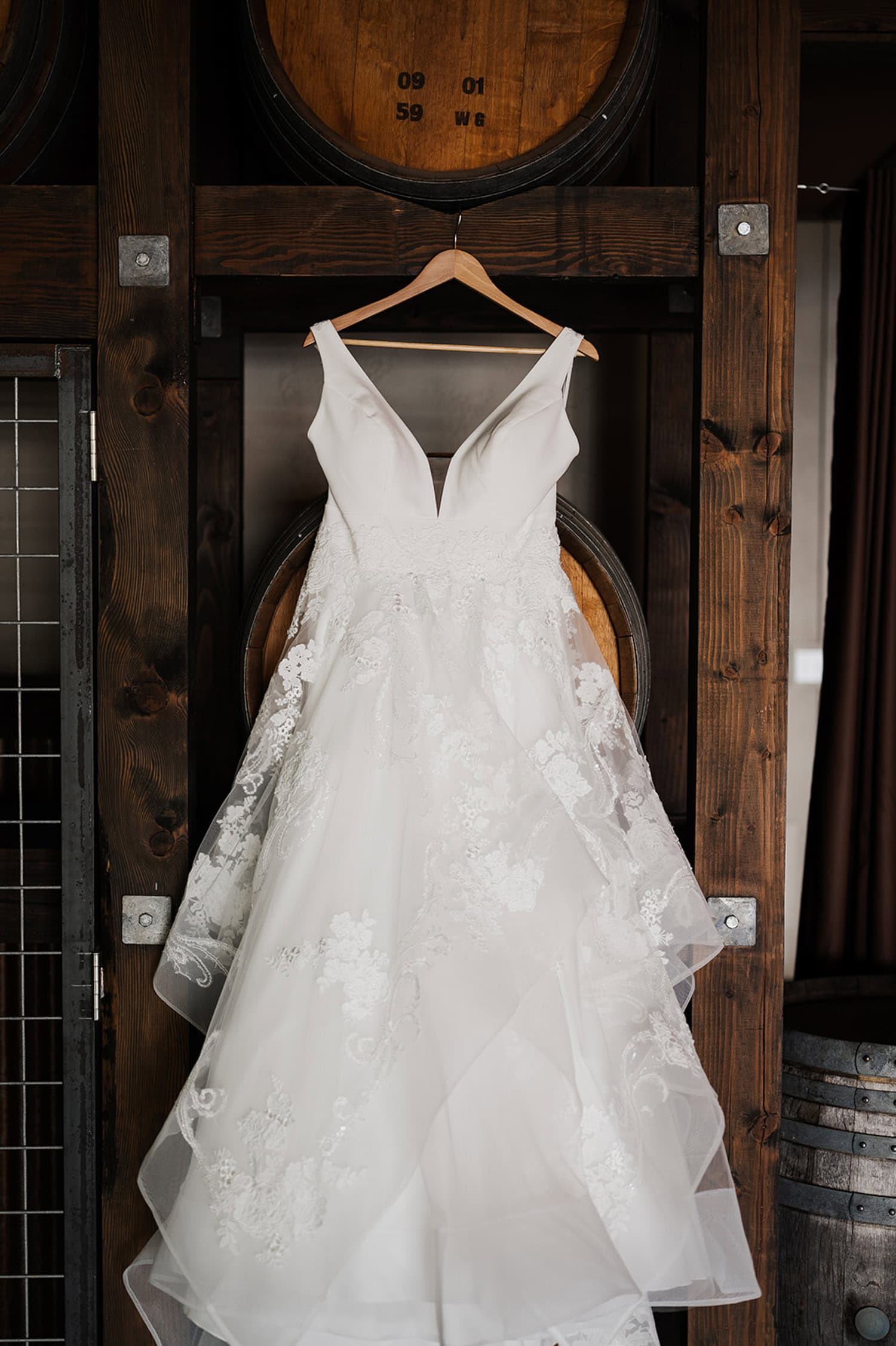 A white wedding dress with lace details hangs in front of large wooden barrels at Gorge Crest Vineyards.