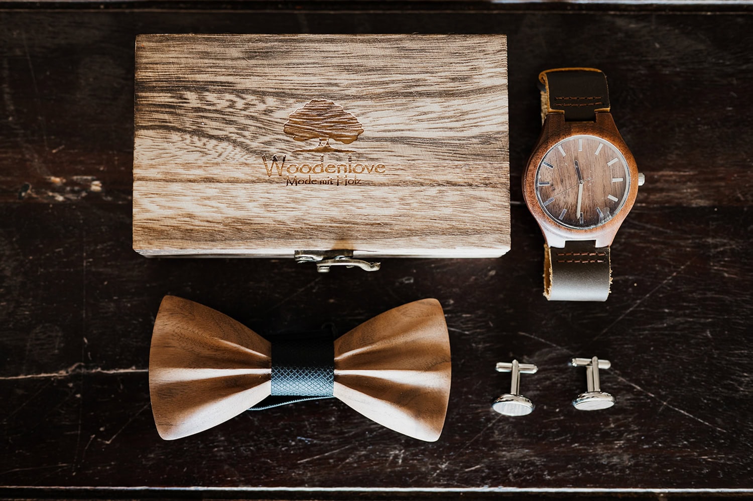 A wooden box with a logo contains a refined wooden watch, bow tie, and cufflinks, placed on a dark wood surface.