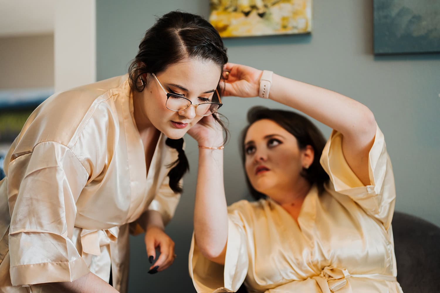 Two women in silk robes are in a cozy room; one is standing with glasses, the other is seated and adjusting her hair. They are preparing for a wedding at Gorge Crest Vineyards.