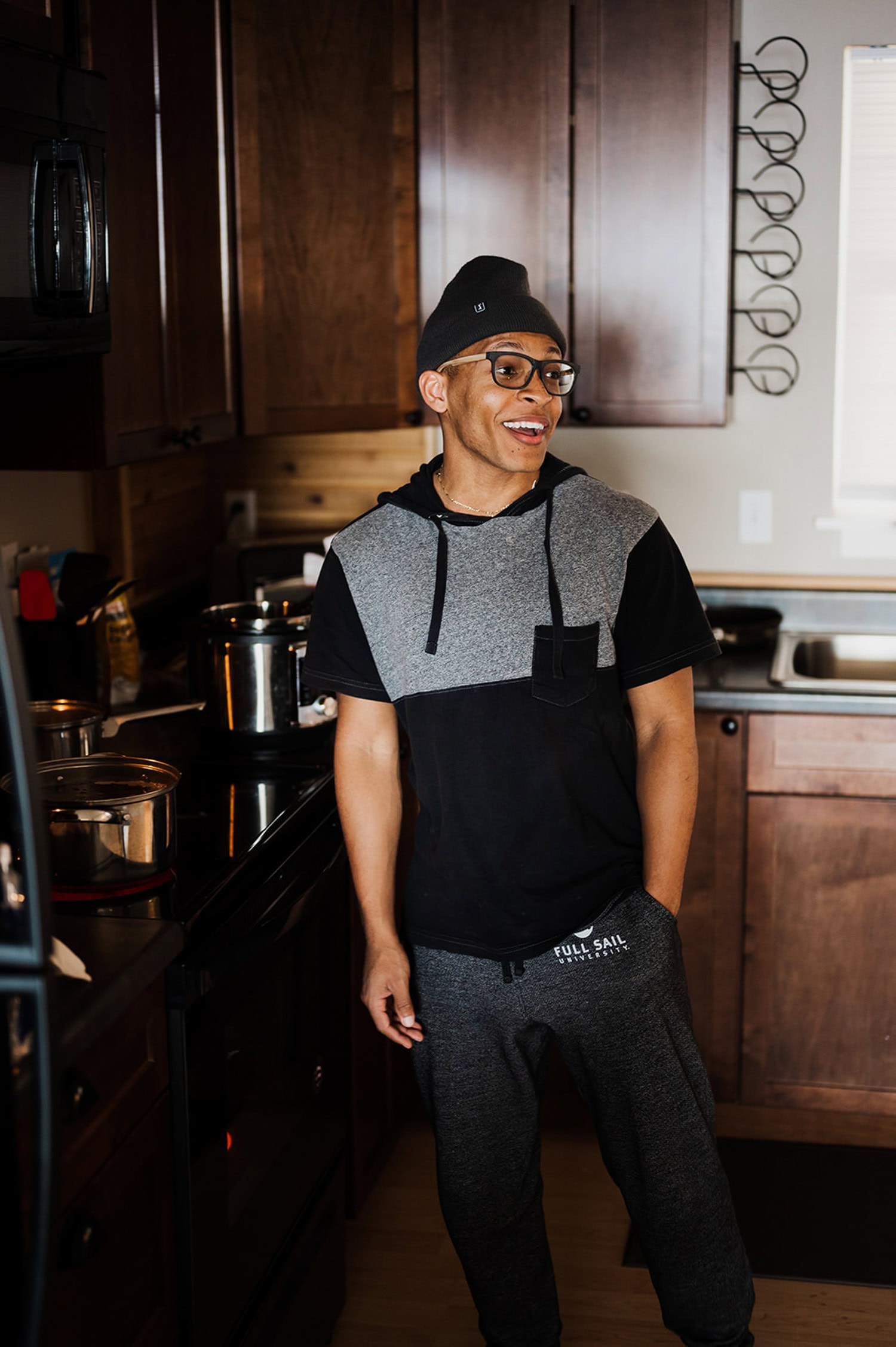 A smiling person in glasses and casual clothes stands near a stove with pots in a cozy kitchen, daydreaming.