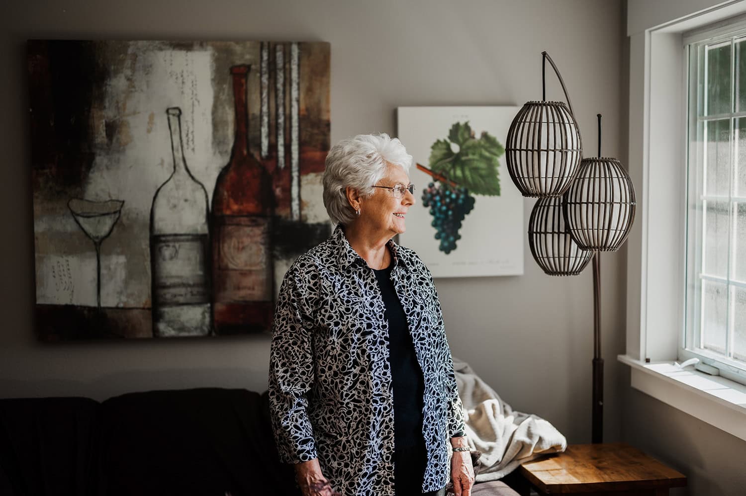 An elderly woman with white hair gazes out the window, standing in a cozy room adorned with wine-themed art.