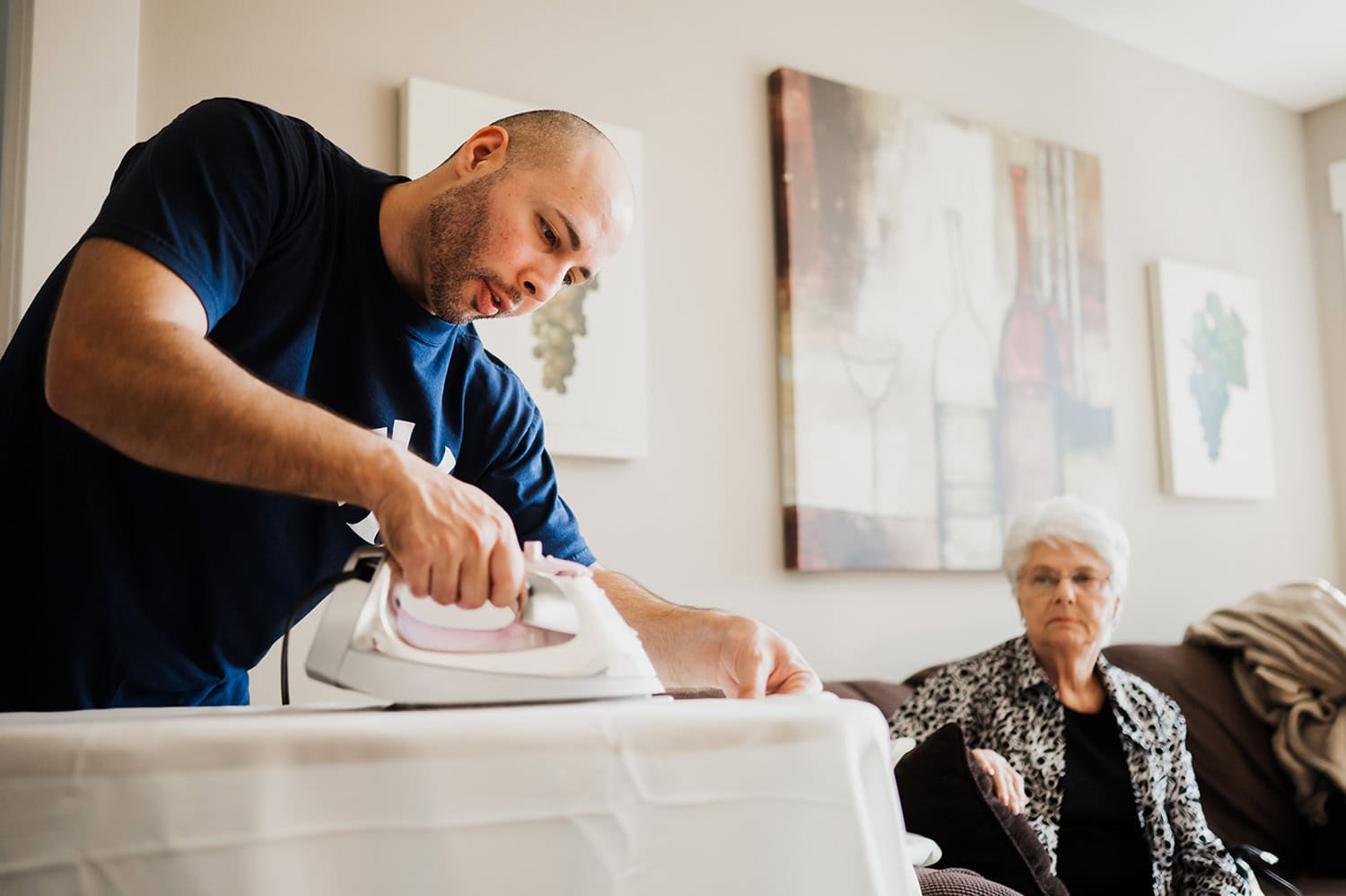 A man carefully irons a shirt while an elderly woman watches from the couch in a cozy living room.