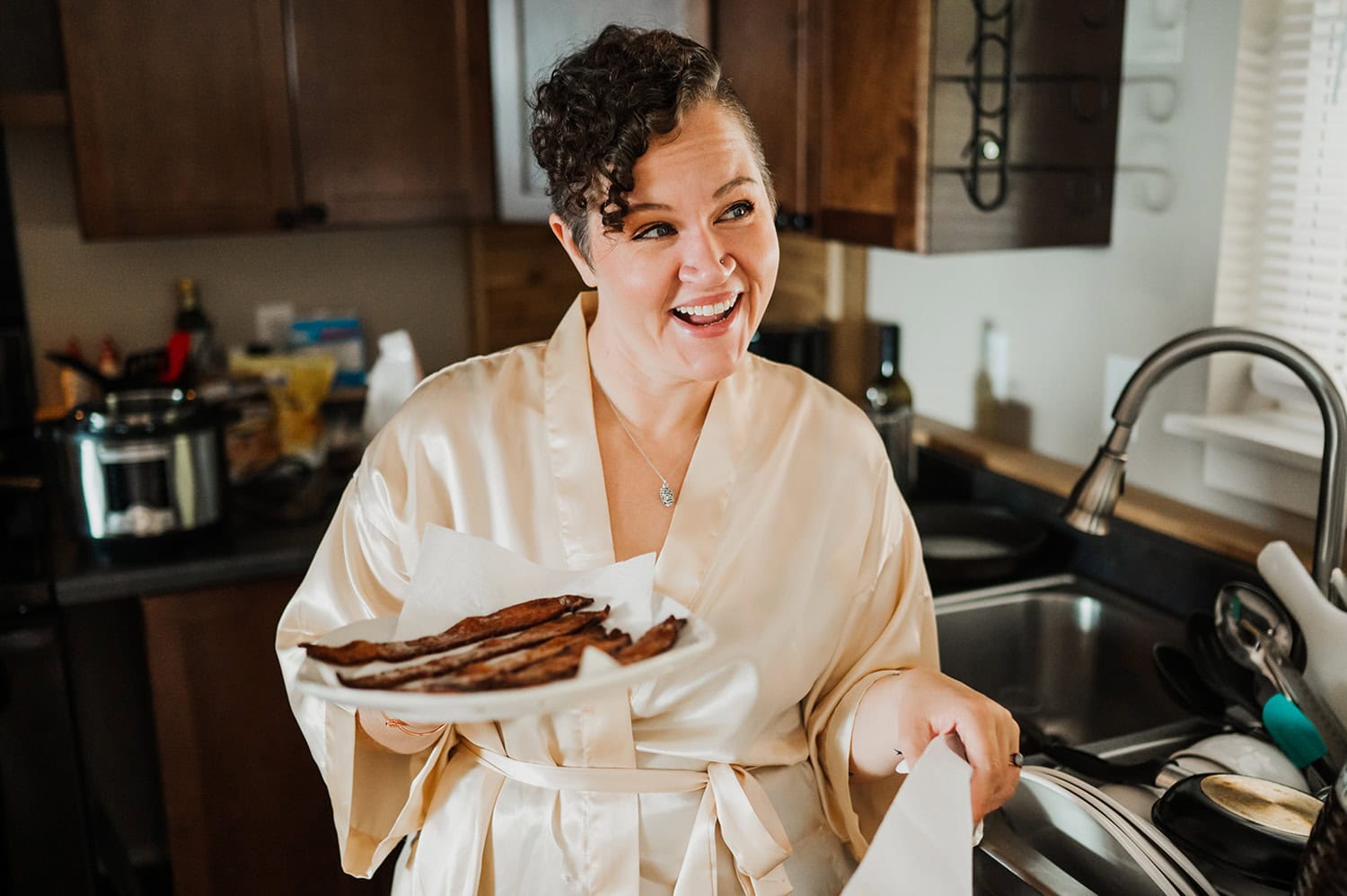 A person wearing a robe stands in a cozy kitchen, smiling while holding a plate of sizzling cooked bacon. The atmosphere suggests a peaceful morning.