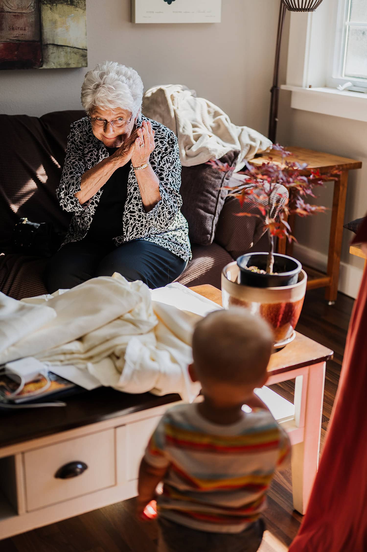 In a cozy living room with a potted plant, an elderly woman makes a playful face at a toddler.