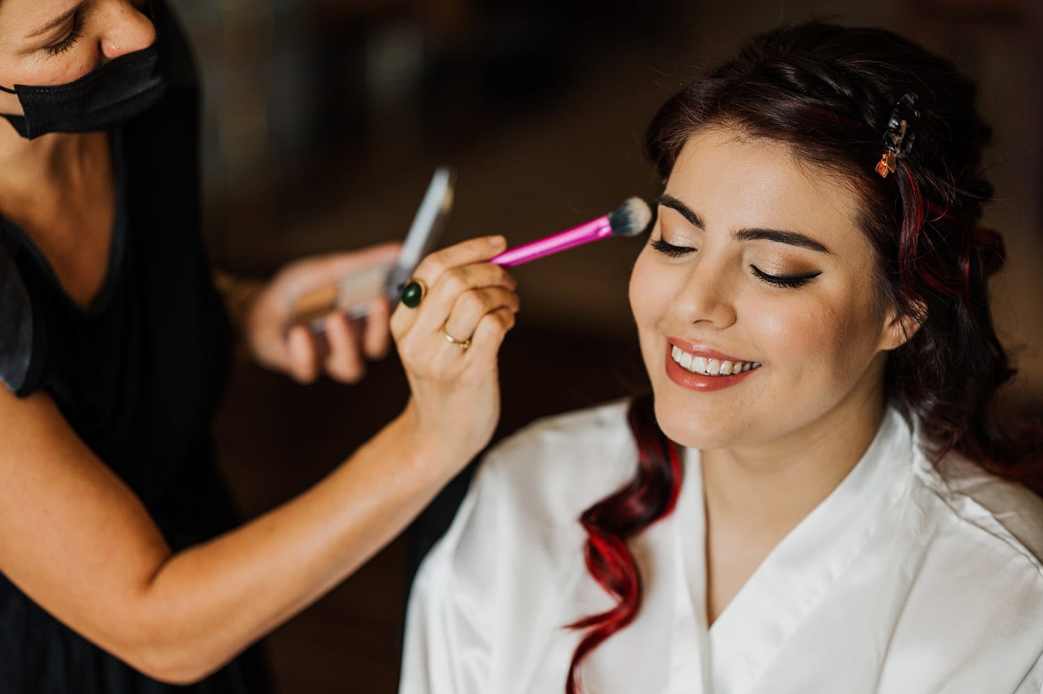 At a Gorge Crest Vineyards wedding, a bride in a white robe smiles brightly while makeup is carefully applied with a pink brush.