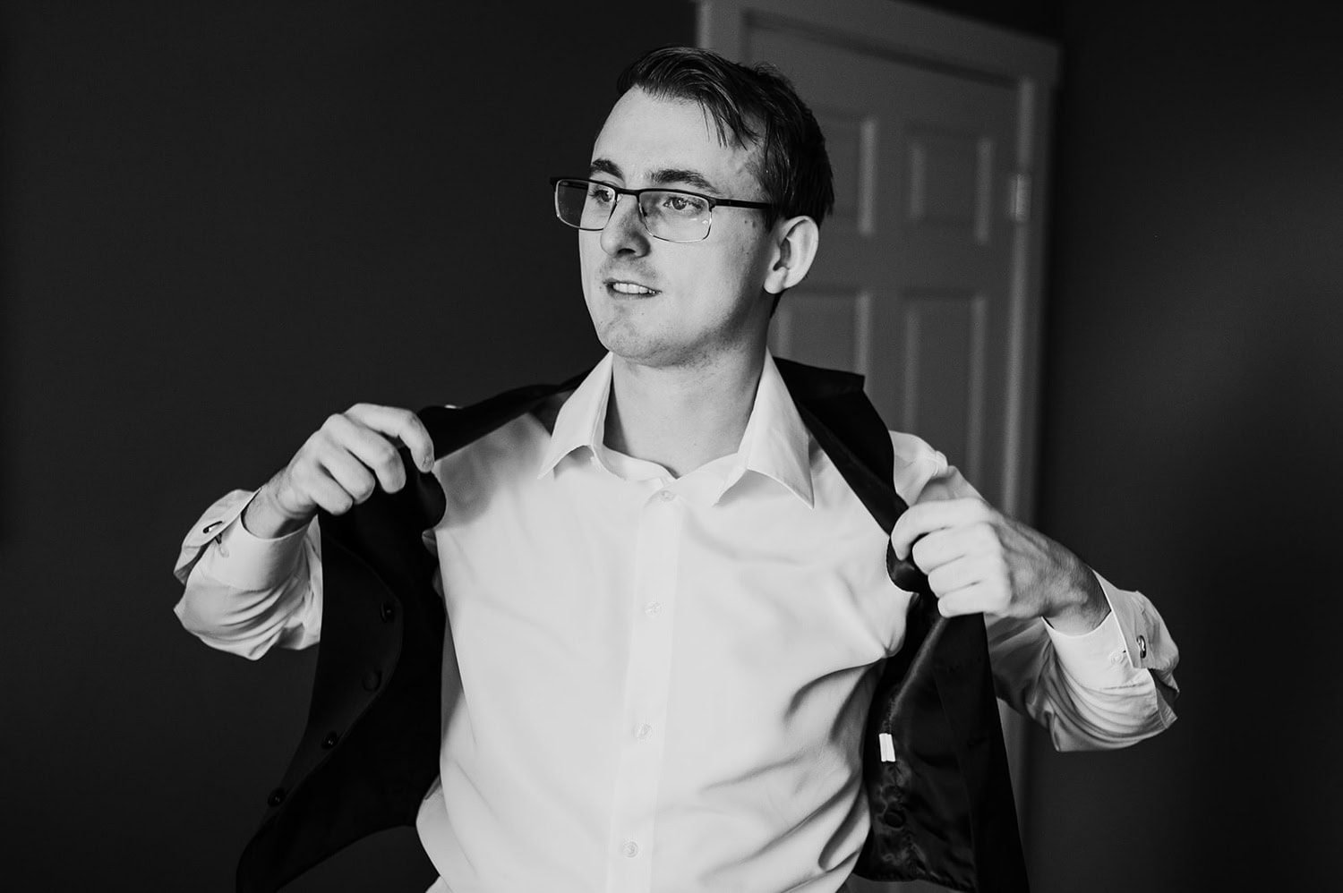 Groom wearing glasses putting on a jacket over a white shirt, standing indoors at Gorge Crest Vineyards in a black and white photo.