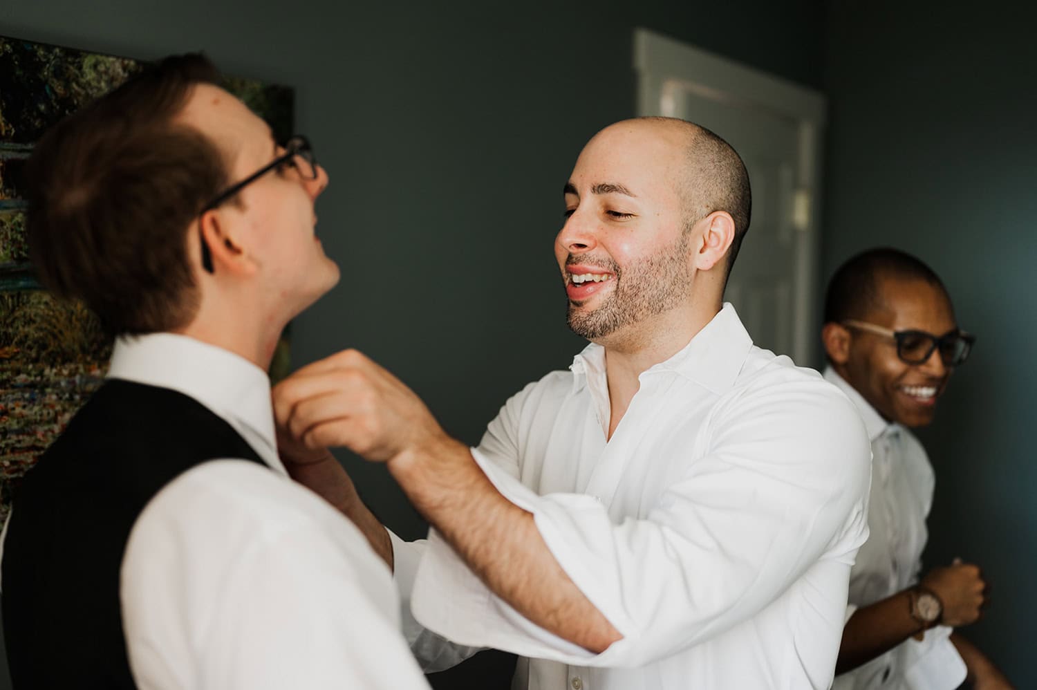 Three men smile warmly as they prepare for the event; one adjusts anothers tie in a sunlit room, with anticipation of a Gorge Crest Vineyards wedding in the air.