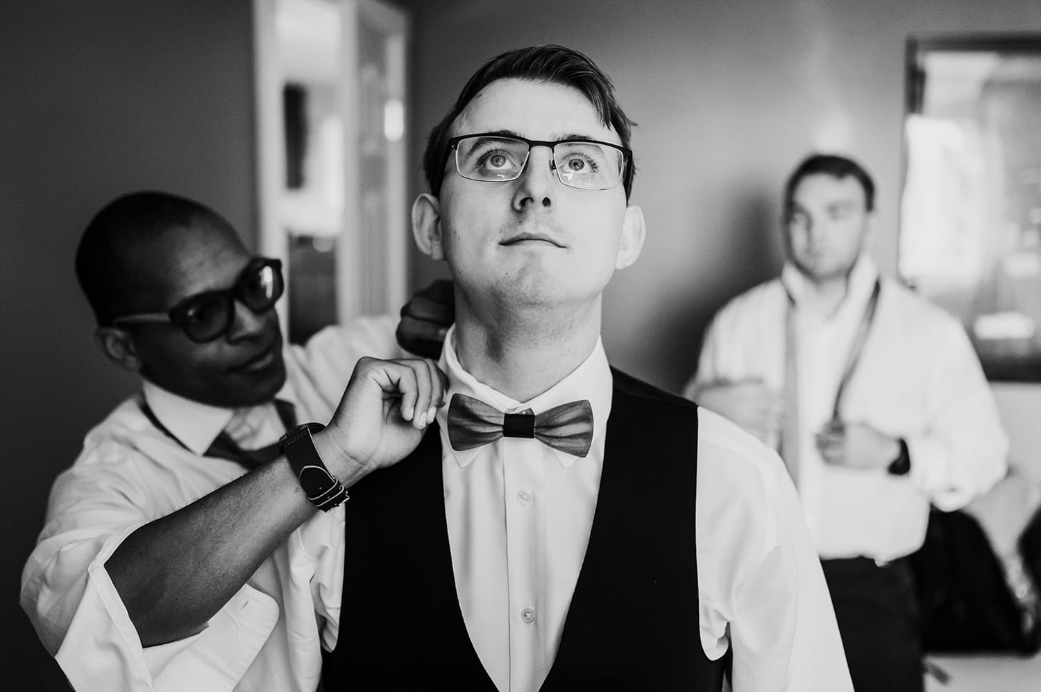 A person adjusts a bow tie for a groom, with a third person in the background getting ready at Gorge Crest Vineyards.