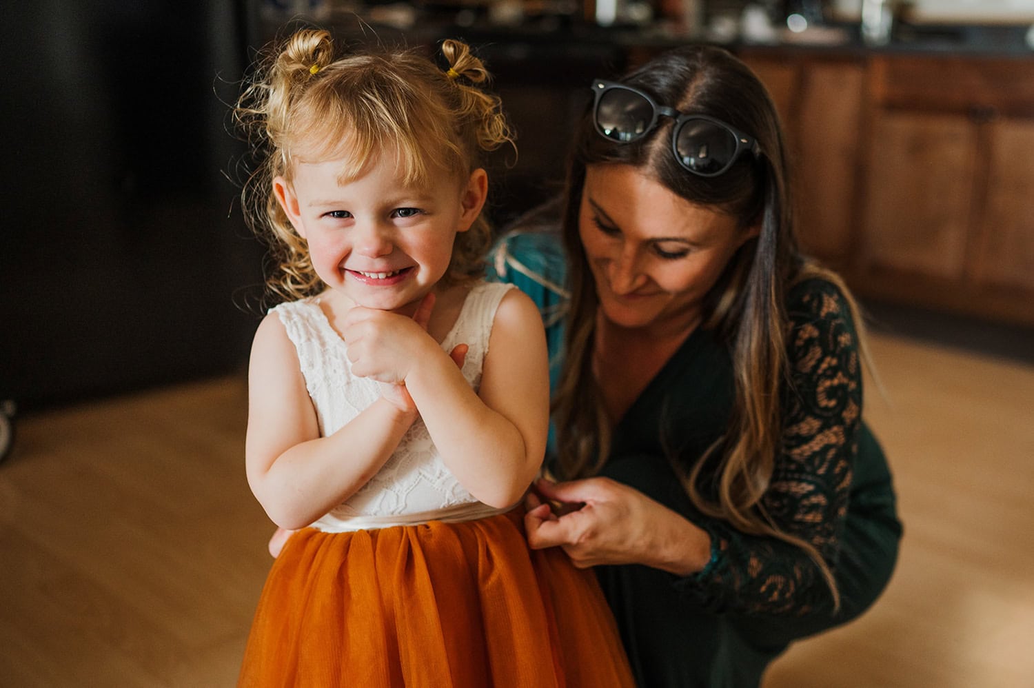 A young girl in a dress stands indoors, smiling as a woman adjusts her outfit.
