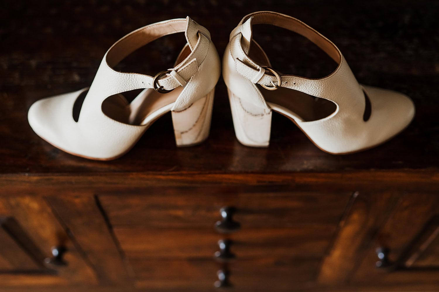 A pair of elegant beige high-heeled shoes with rounded cutouts and buckle straps placed on a wooden surface.