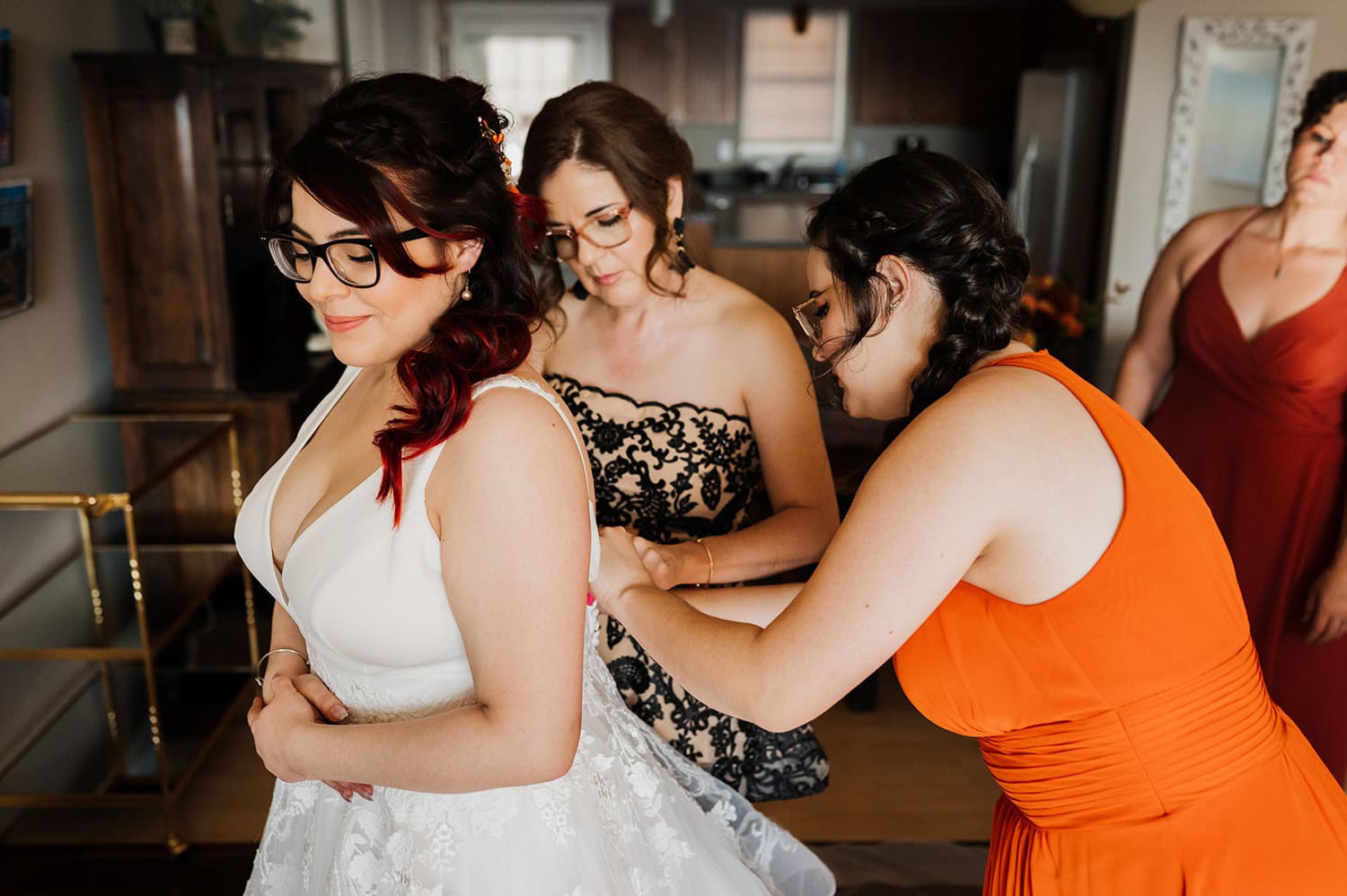 A bride dressed in white with glasses stands in a room at Gorge Crest Vineyards, surrounded by bridesmaids who are assisting her.