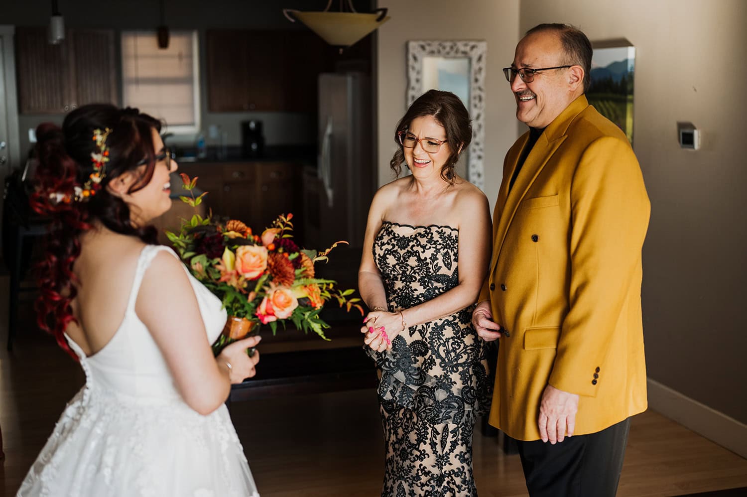 In a cozy home, the bride holds her bouquet while two guests in elegant formal attire smile at her.
