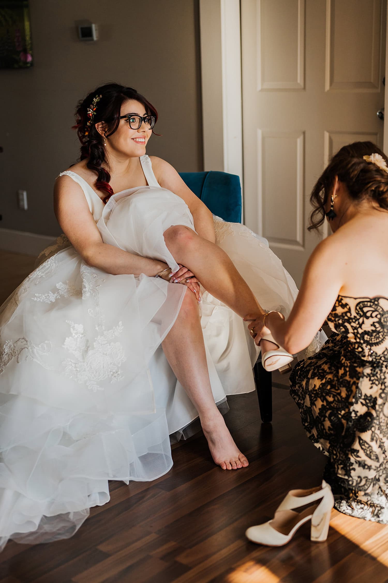 At Gorge Crest Vineyards, the bride sits smiling while another woman helps her with a shoe. Her elegant wedding dress drapes gracefully over her leg. The scene is set against a vineyard backdrop with lush green hills and clear skies.