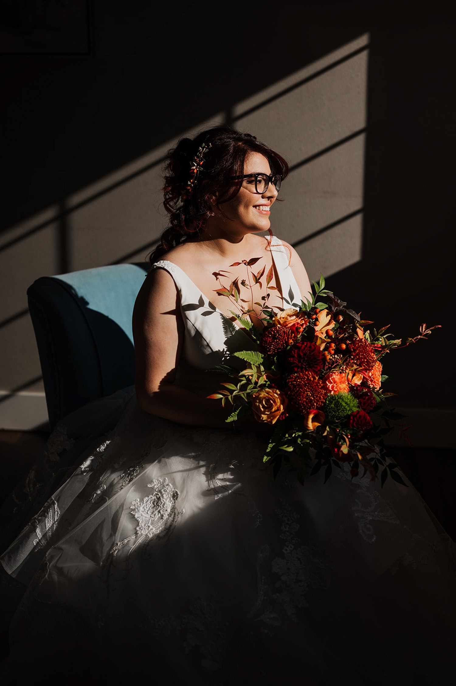 A bride in a white gown holds a colorful bouquet while sitting on a blue chair with sunlight streaming through window blinds at Gorge Crest Vineyards.