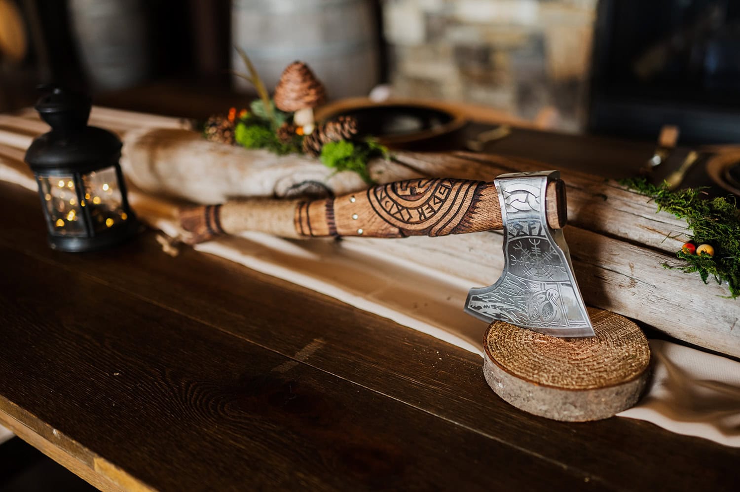 An ornate decorative axe with detailed designs is placed on a wooden table next to a small lantern and a pine cone.