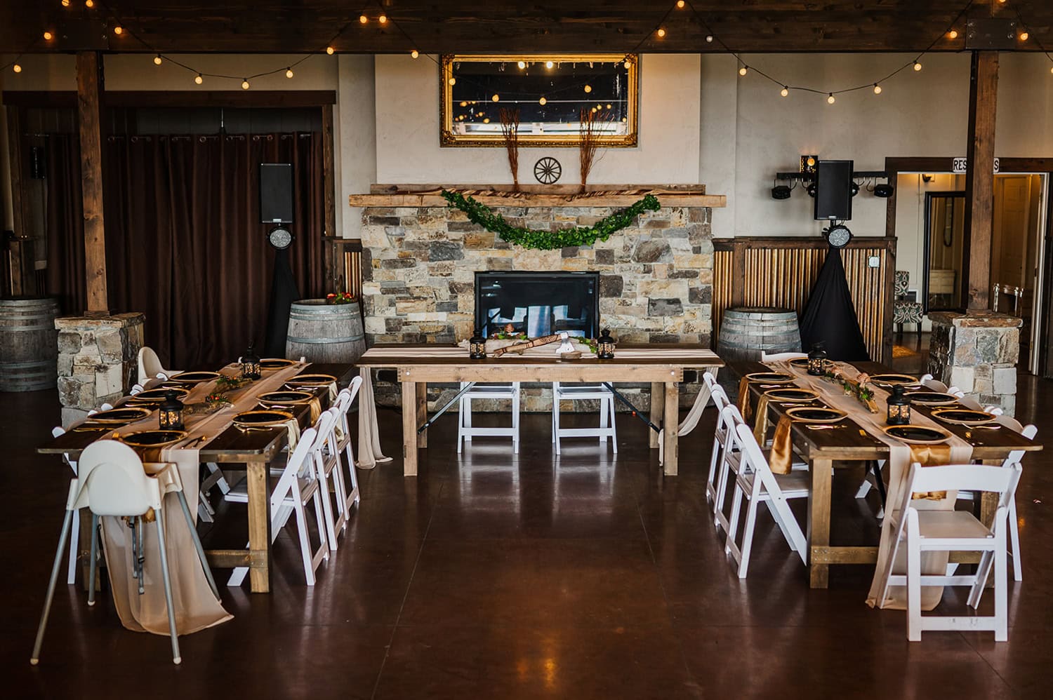 A wedding reception at Gorge Crest Vineyards with rustic wooden tables, white chairs, and twinkling string lights set against a stone fireplace backdrop, creating a warm and inviting atmosphere.
