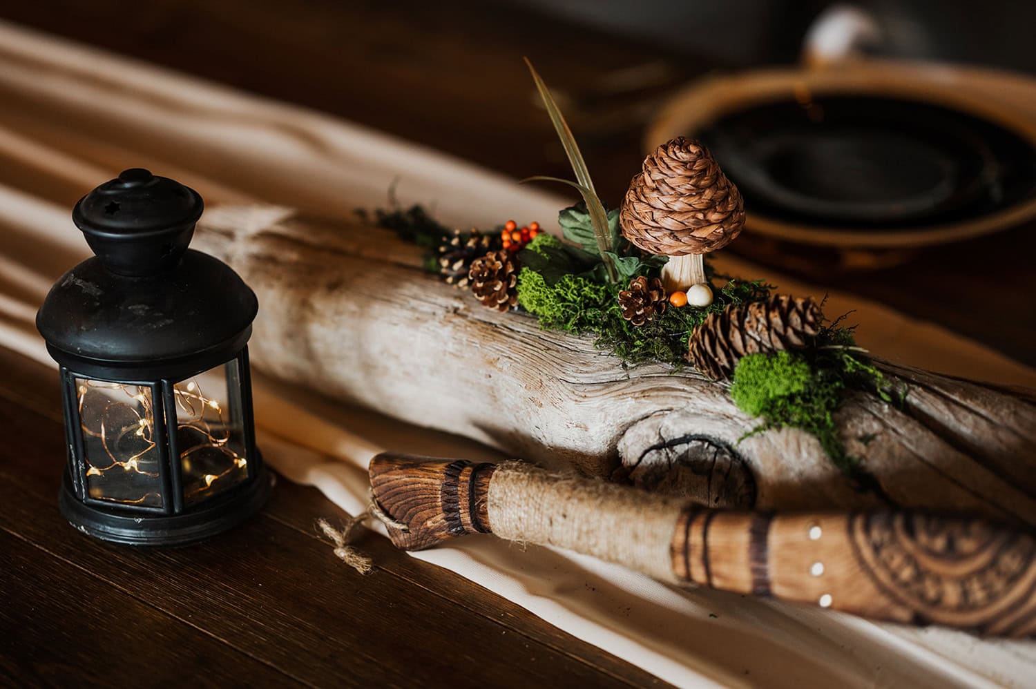 A wooden log adorned with pinecones, mushrooms, moss, and a black lantern is arranged on a table.