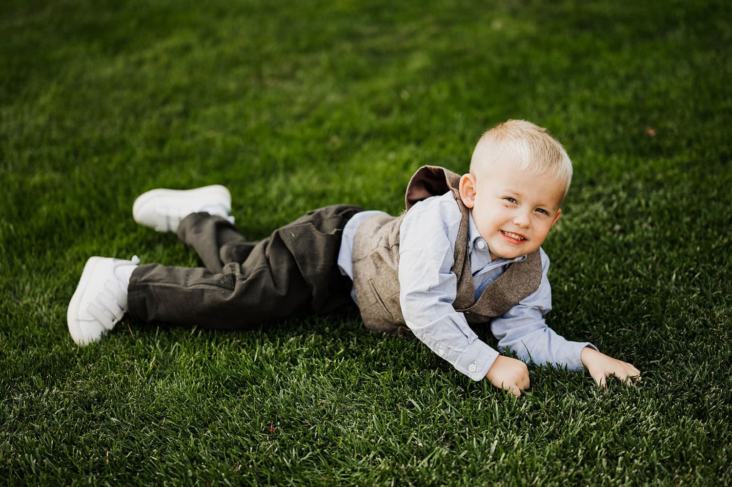 A young boy in a vest and tie lies on lush green grass, smiling with satisfaction. Behind him is the peaceful scenery of Gorge Crest Vineyards.