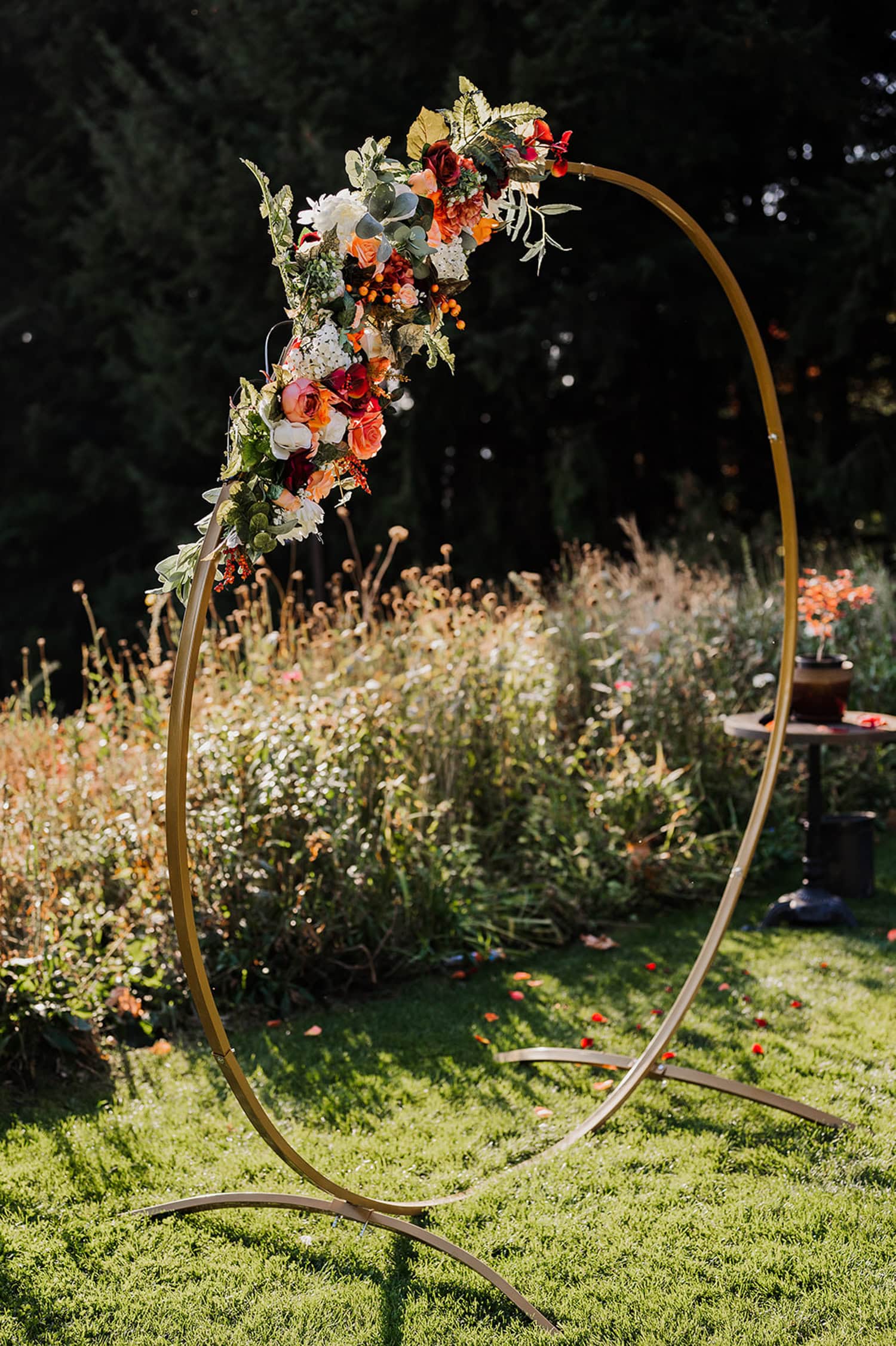 A round gold wedding arch adorned with colorful flowers, set outdoors on a grassy area at Gorge Crest Vineyards, with lush greenery in the background.