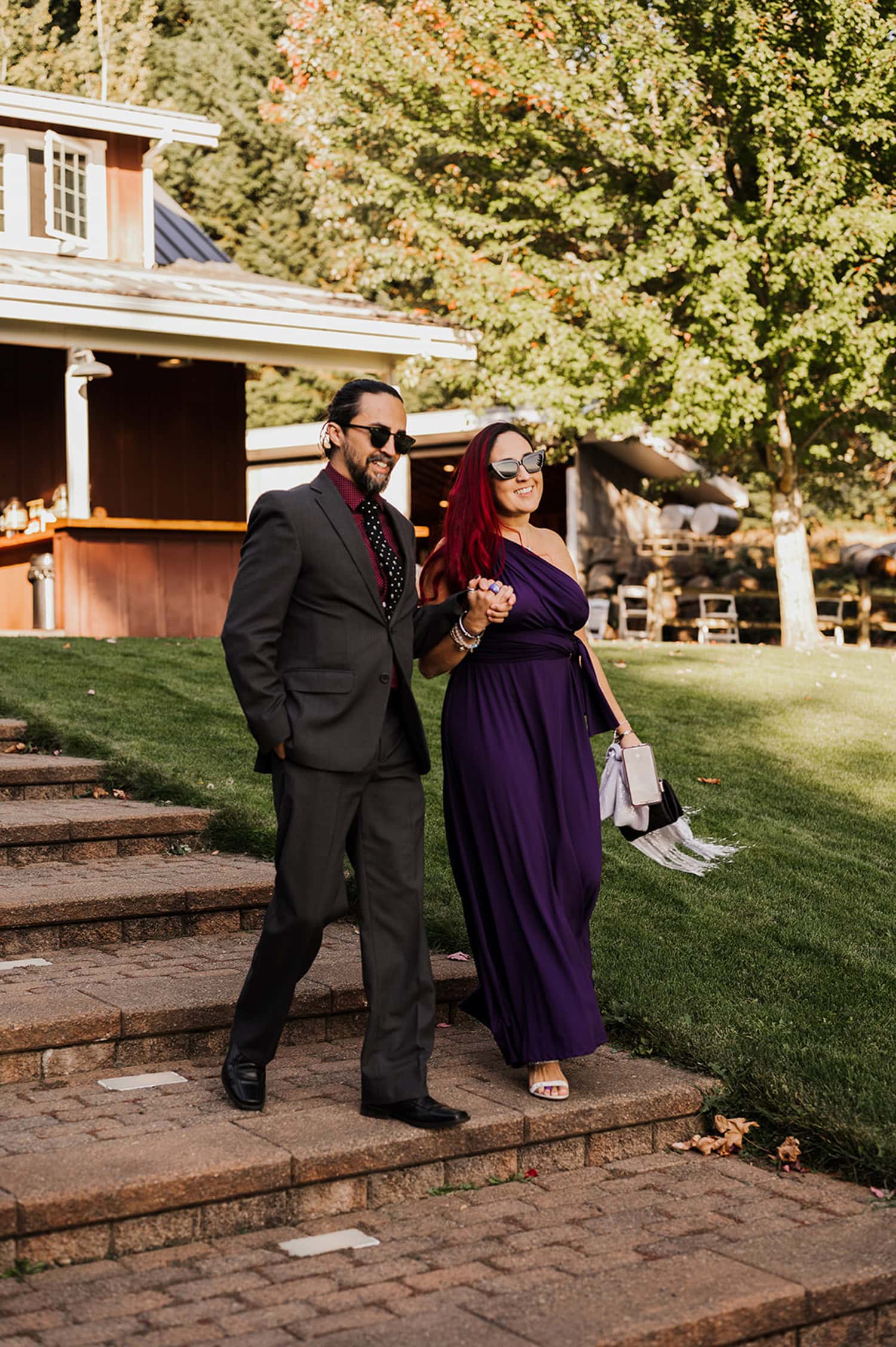 A couple in formal attire descends brick steps outdoors on a sunny day, with lush trees in the background at their Gorge Crest Vineyards wedding.