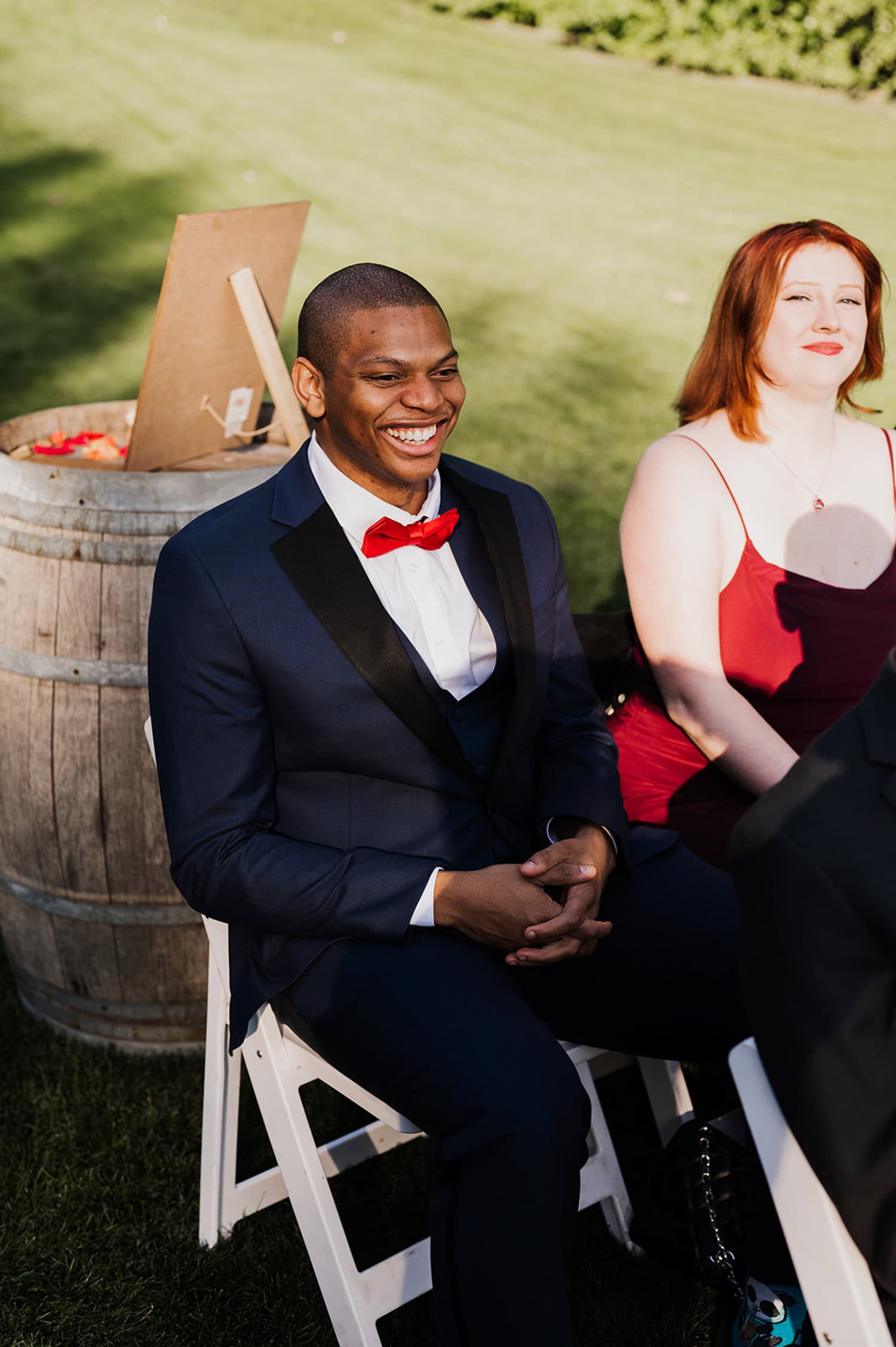 A man in a blue suit with a red bow tie sits next to a woman in a red dress at Gorge Crest Vineyards. Both are smiling and surrounded by the lively atmosphere of the wedding celebration.