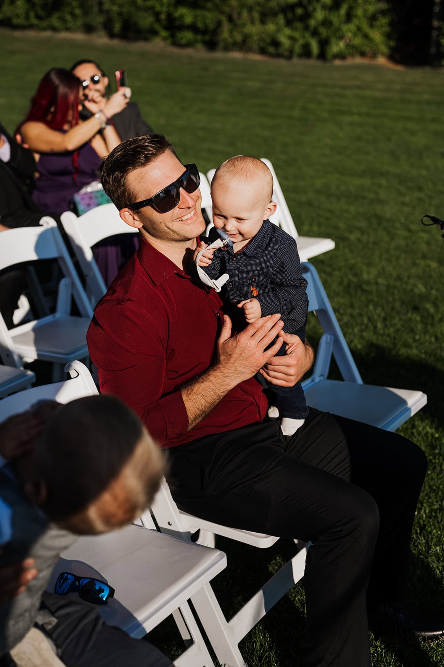 Man in sunglasses holding a smiling baby while seated outdoors on white chairs, with people mingling in the background at Gorge Crest Vineyards.