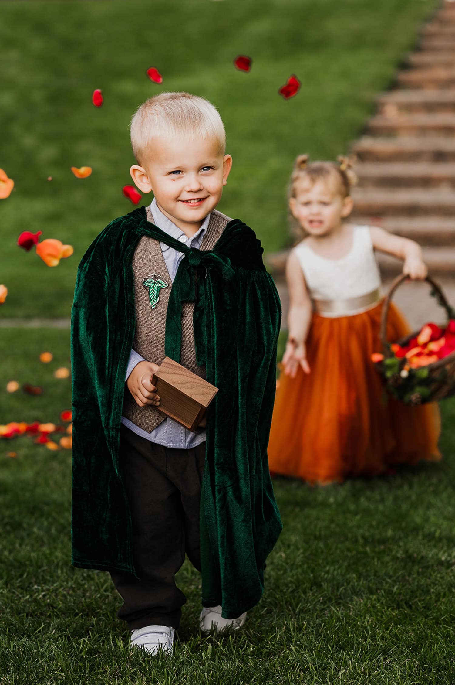 A smiling boy in a green cape holds a wooden box while walking down a grassy path at Gorge Crest Vineyards, with a girl behind him gently sprinkling flower petals.