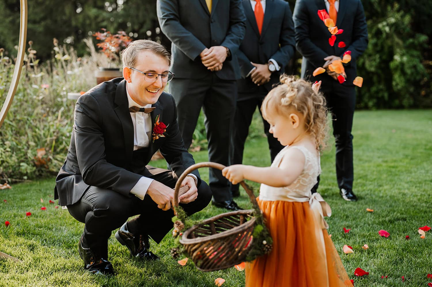 A man in a suit kneels on the grass at Gorge Crest Vineyards, smiling at a young girl holding a basket with flower petals around them.