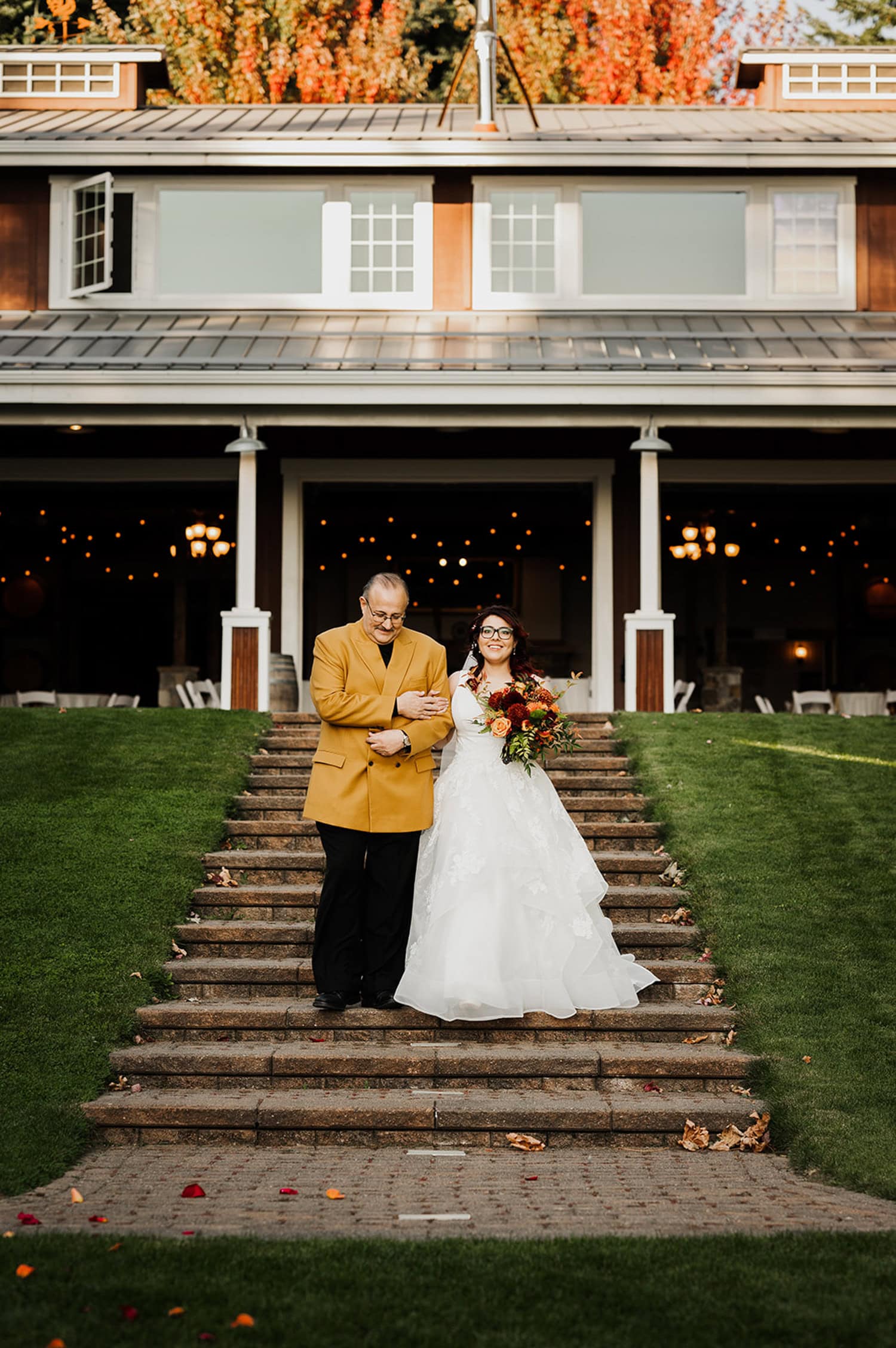 A bride in a white dress walks arm-in-arm with a man in a yellow jacket down outdoor steps at Gorge Crest Vineyards, with a building visible in the background.