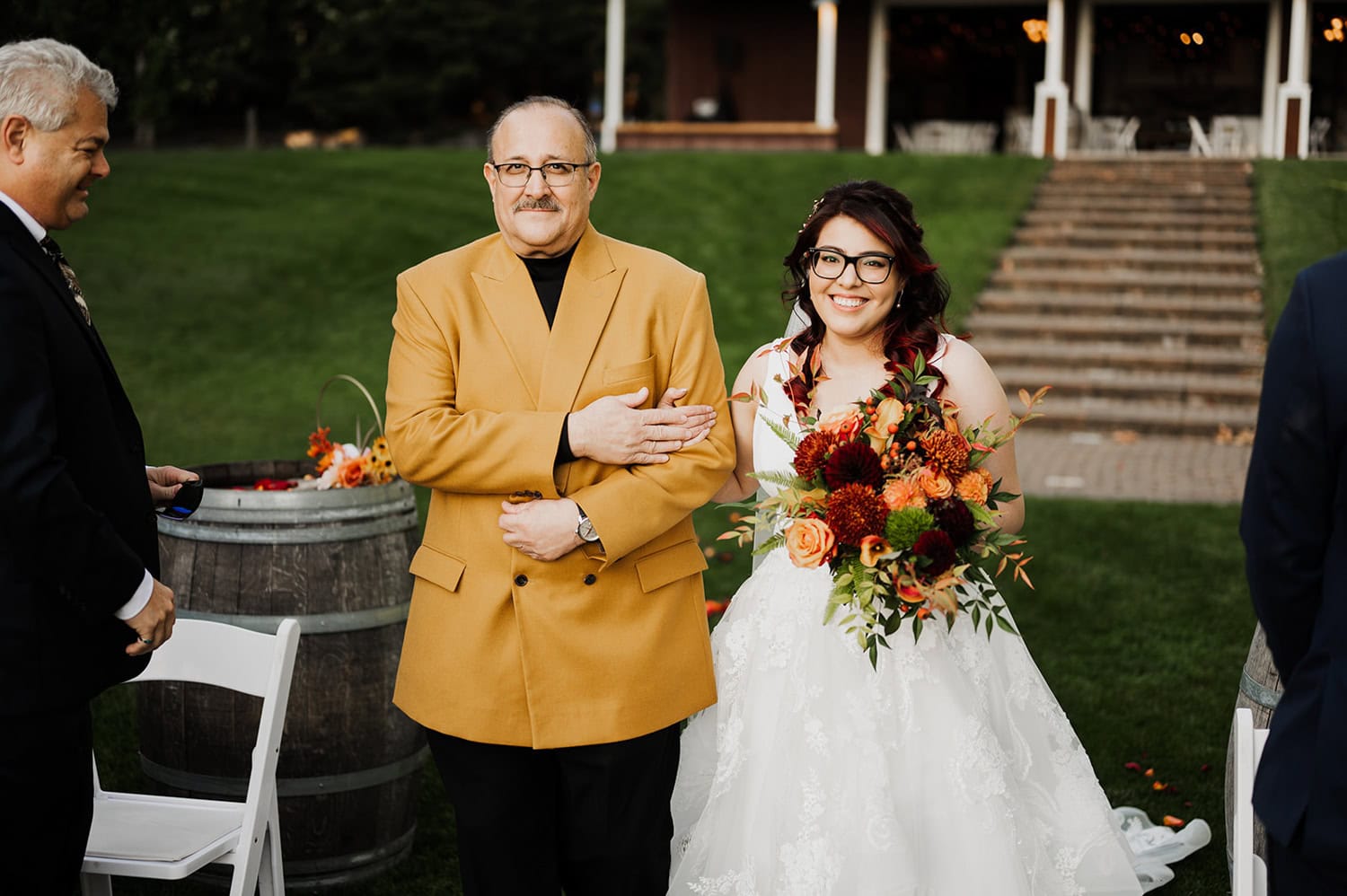 At Gorge Crest Vineyards, a bride in a white gown holds a vibrant bouquet and walks down the aisle with a man in a yellow jacket during an outdoor ceremony.