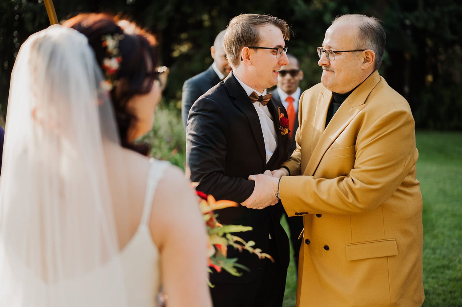 A groom shakes hands with a man in a tan suit while the bride stands nearby at Gorge Crest Vineyards, surrounded by the tranquil outdoor scenery.