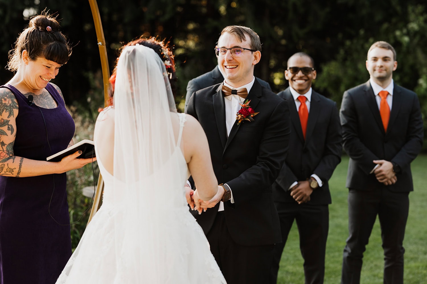 A couple exchanges vows outdoors at Gorge Crest Vineyards, with a smiling officiant and groomsmen in suits looking on against a backdrop of vineyards and mountains.