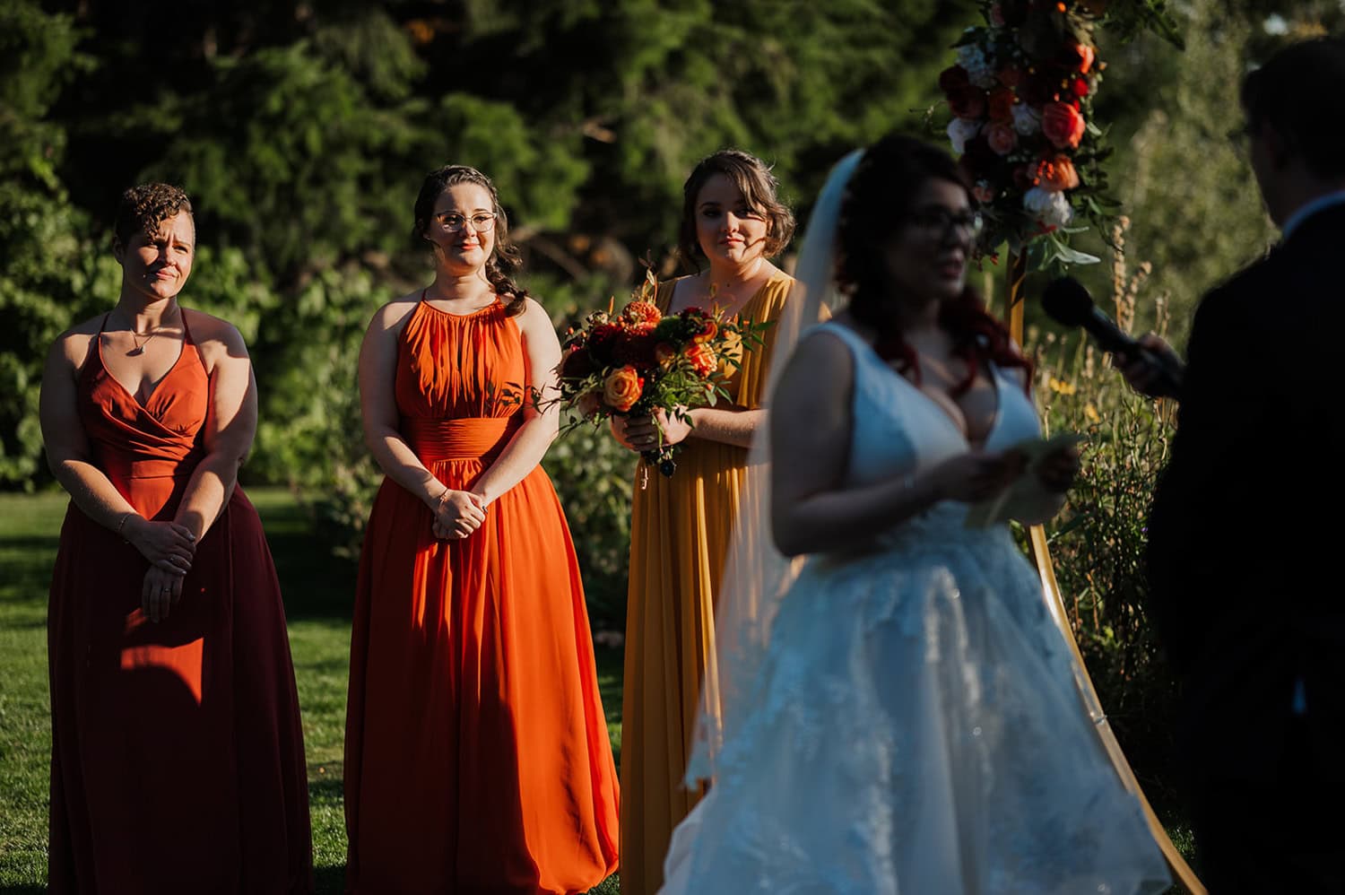 A bride speaks to her guests in an outdoor setting at Gorge Crest Vineyards, accompanied by three bridesmaids in bright red and orange dresses.