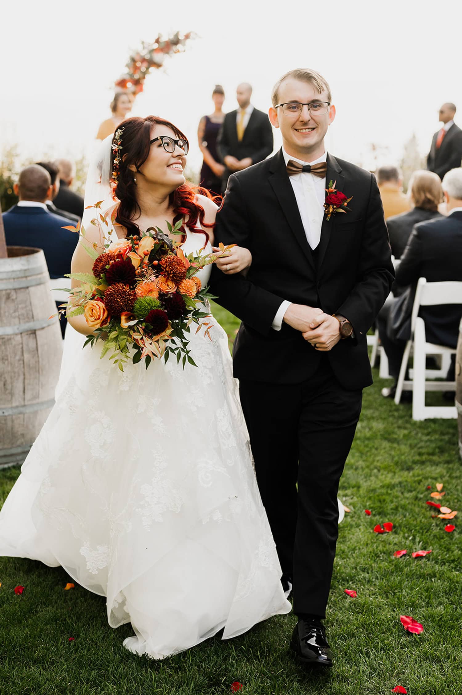 A bride and groom walk arm in arm down an outdoor aisle at Gorge Crest Vineyards, smiling with an autumn-themed floral arrangement surrounding them.