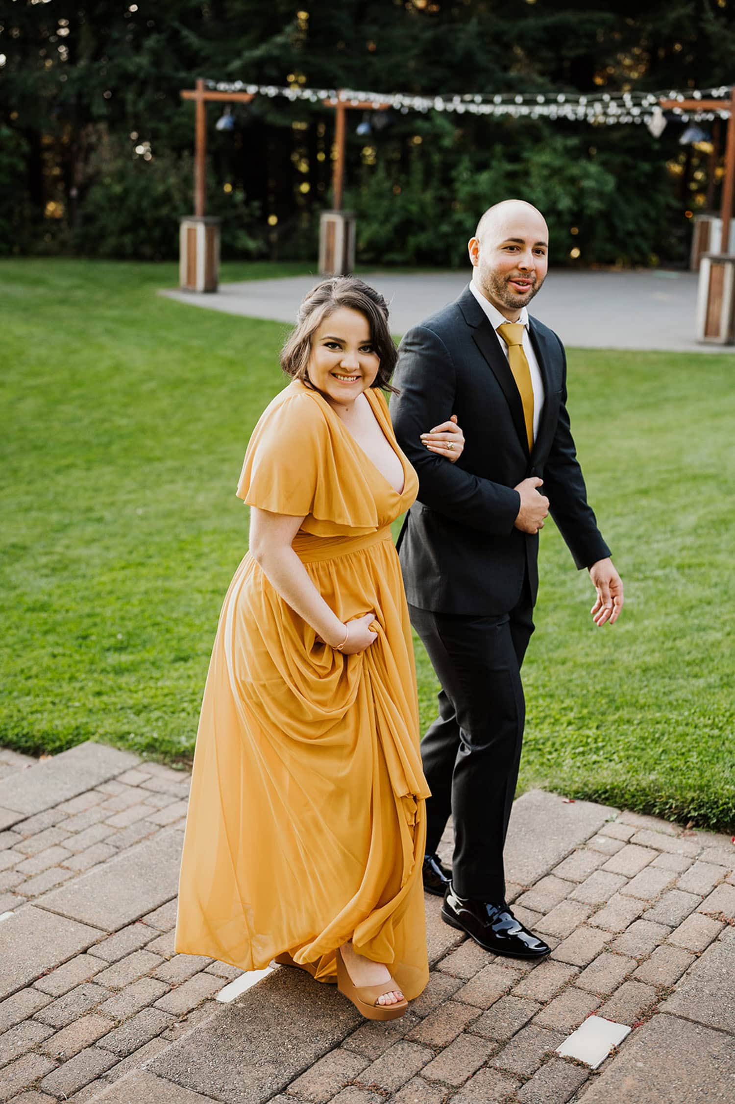 A woman in a yellow dress and a man in a suit walk along a path at Gorge Crest Vineyards, smiling joyfully. Lush green lawns and trees surround them in the background.